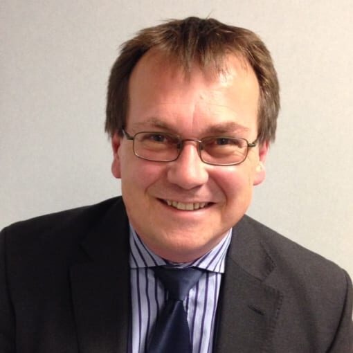 Clive Nelson in dark suit, striped shirt and blue tie smiles at the camera