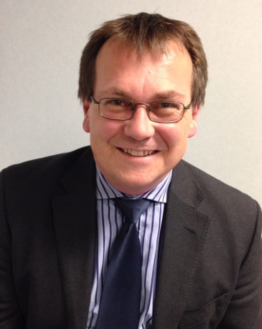 Clive Nelson in dark suit, striped shirt and blue tie smiles at the camera