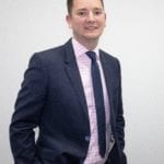 Daniel Hickman in a blue suit, shirt and tie stands with his hands in his pockets.
