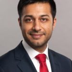 Manroop Khela in a dark suit and red tie looks at the camera