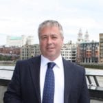 Mark Summerfield stands by a river with a city skyline in the distance