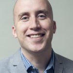 Philip wearing a grey blazer and blue check shirt, smiles towards the camera