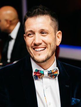 Daniel wearing a black blazer and colourful bow tie, laughs in front of the camera