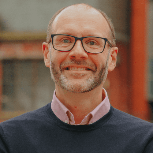 Alan McBride against a blurred background, wearing glasses smiling at camera.