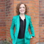 Lisa Mcllvenna wearing a green blazer against a red brick wall as background.