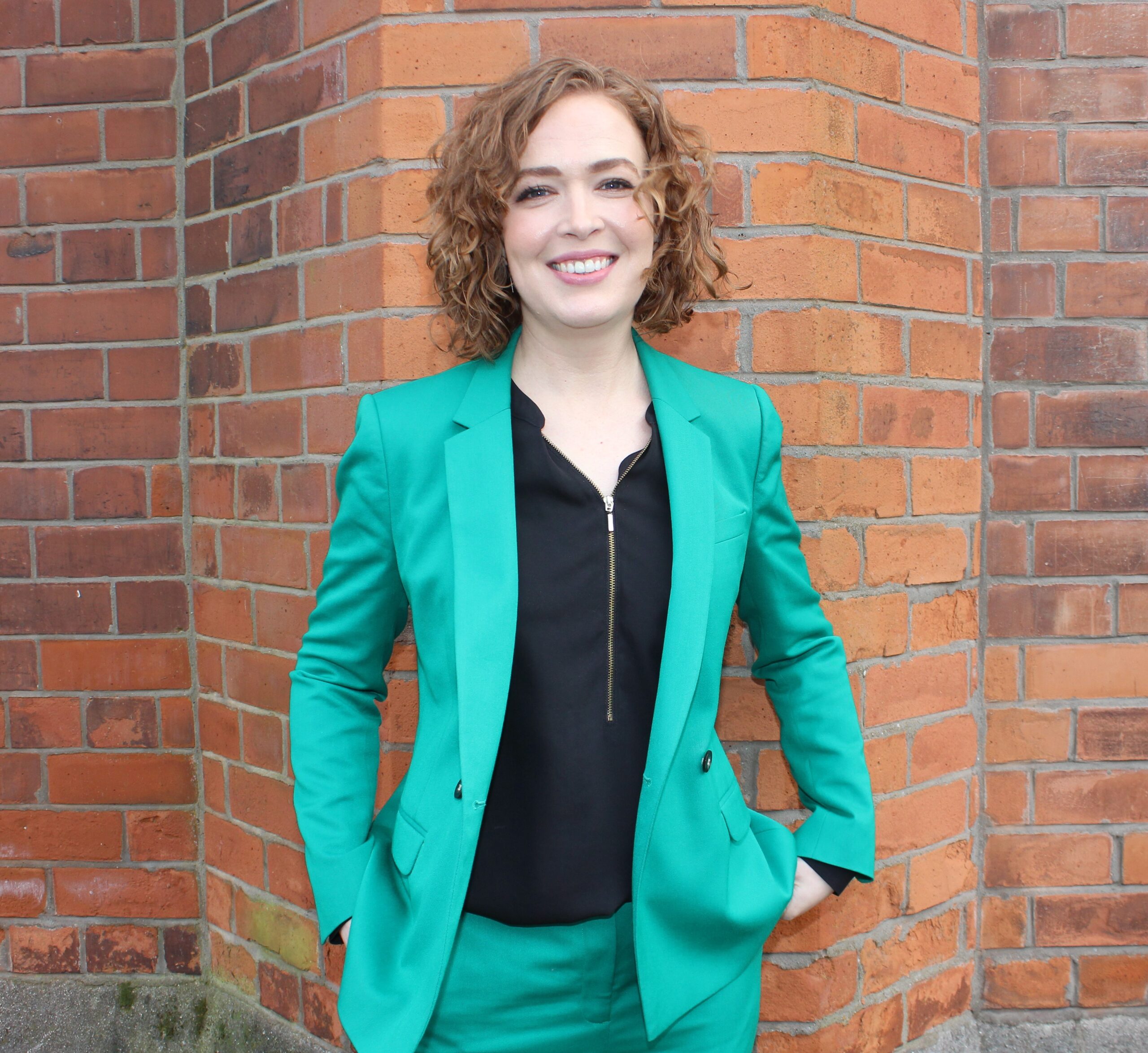 Lisa Mcllvenna wearing a green blazer against a red brick wall as background.